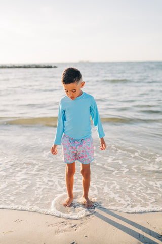 Child standing in shallow water at the beach wearing a light blue long-sleeve shirt and patterned shorts.
