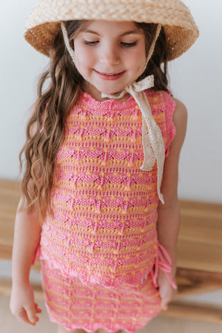 Young girl wearing a pink and yellow striped dress with a straw hat indoors.