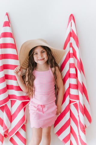 Young girl in a pink checkered dress and sun hat standing between two red and white striped towels.