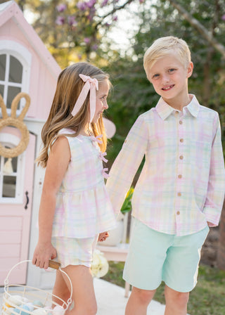 Two children, a girl and a boy, standing outdoors with a pink playhouse in the background.