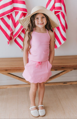 Young girl in a pink gingham athletic set and sun hat standing in front of red and white striped towels.