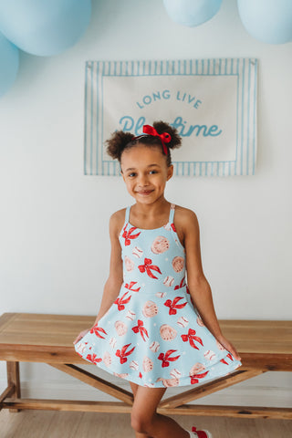 Young girl in a light blue dress with red patterns sitting on a wooden bench.