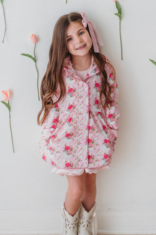 Young girl in a floral rain jacket standing against a wall with pink tulips.