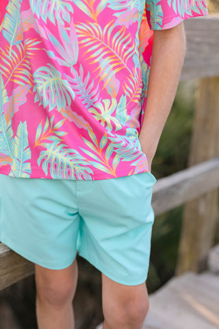 Person wearing a bright pink shirt with tropical leaf pattern and light blue shorts, sitting on a wooden bench.