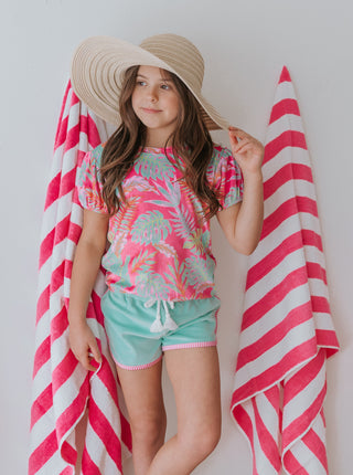 Young girl in a colorful outfit with a straw hat and pink and white striped towel on a plain background