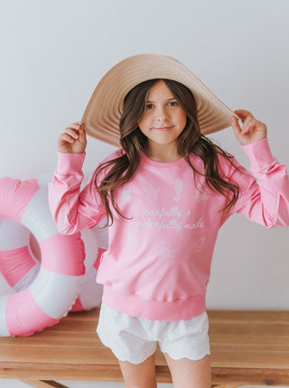 Child wearing a pink sweatshirt with text, white shorts, and a wide-brimmed hat, standing next to a pink inflatable ring.