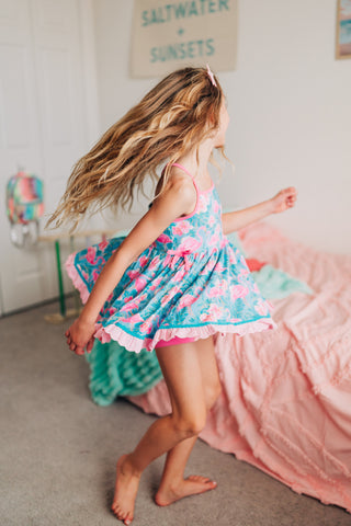 Young girl in a colorful dress dancing in a bedroom with pink bedding and a 'Saltwater Sunsets' sign.