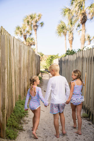 Three children in matching outfits walking along a sandy path with palm trees in the background.