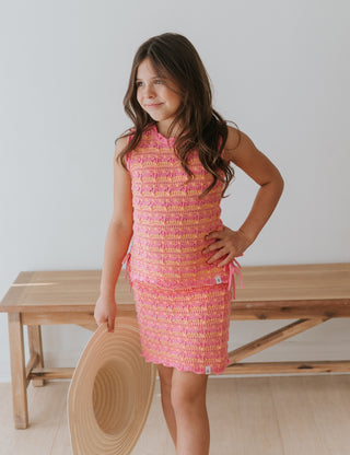 Young girl in a pink dress standing next to a wooden bench holding a hat.