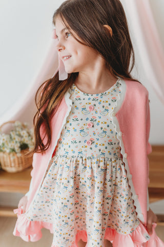 Young girl wearing a floral dress with a pink cardigan in a bright room.