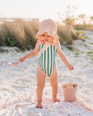 Child in a striped swimsuit and sun hat standing on a sandy beach with a bucket and shovel.