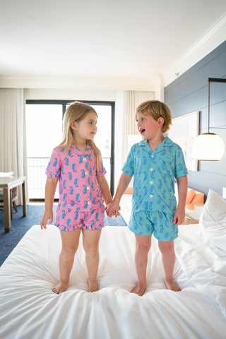 Two children in matching pajamas standing on a bed in a room with a window and lamp.