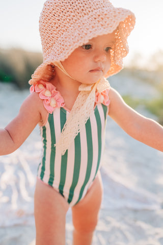 Child wearing a green and white striped swimsuit with a pink sun hat on a beach.