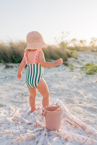 Child in a green and white striped swimsuit and straw hat on a sandy beach.