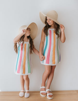 Two young girls wearing colorful striped dresses and hats against a white wall.