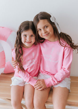 Two young girls wearing pink long-sleeve shirts with a graphic design, sitting on a wooden floor.