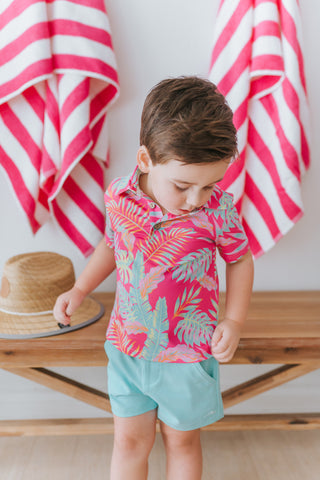 Child wearing a pink tropical shirt and light blue shorts standing in front of pink and white striped towels.