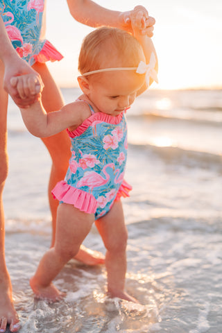Baby in a colorful swimsuit with flamingo design standing in shallow water at the beach.