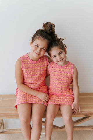 Two young girls in matching pink outfits sitting on a wooden bench.