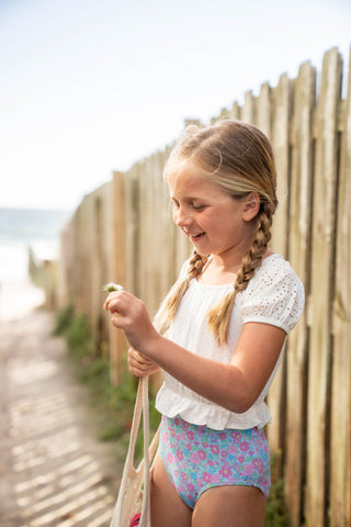 Young girl in a swimsuit standing on a wooden boardwalk with a wooden fence and beach in the background.