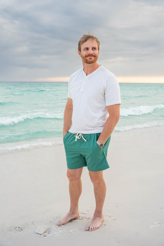 Man standing on a beach wearing a white shirt and green shorts with a cloudy sky and ocean in the background.