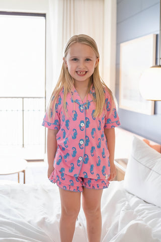 Young girl in a pink pajama dress standing on a bed in a bright bedroom.