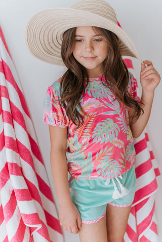 Young girl wearing a pink and green palm leaf-patterned top and light aqua shorts, standing against a white background with a pink and white striped towel.