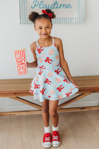 Young girl wearing a light blue dress with Red bows, baseballs, and baseball glove pattern, standing indoors.