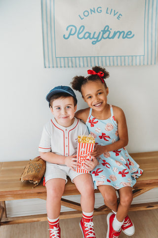 Two children sitting on a bench with popcorn, smiling at the camera, with a sign that reads 'Long Live Playtime' in the background.