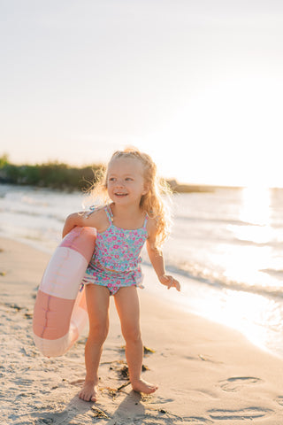 Young girl in a swimsuit holding a beach ball on a sandy beach with water in the background.