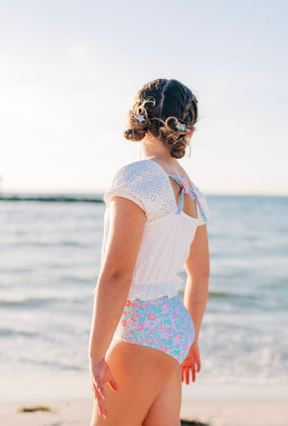 Child in a white lace top and colorful swimsuit standing on a beach.