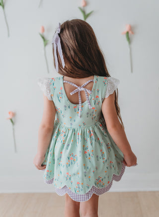 Young girl wearing a light green floral dress with a white bow in her hair, standing against a light background.