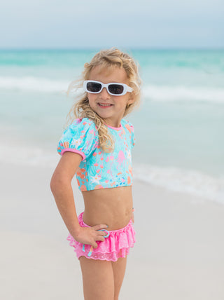 Young girl in a colorful swimsuit and sunglasses standing on a beach.