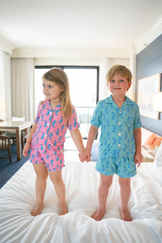 Two children holding hands on a bed wearing pajamas in a bright room.