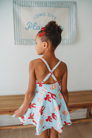 Young girl wearing a light blue dress with Red bows, baseballs, and baseball glove pattern, standing indoors.
