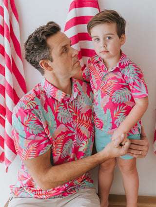 Man and child wearing matching pink tropical shirts in front of red and white striped curtains.