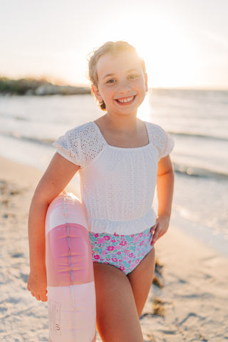 Young girl in a white top and floral swimsuit holding a pink floatie on a beach.
