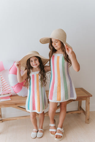 Two young girls wearing colorful striped dresses and hats on a wooden bench.