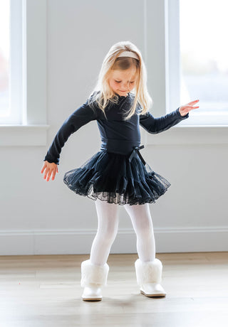 Young girl in a black outfit with a tutu standing in a room with white walls and windows.