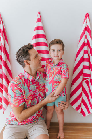 Man and child in matching colorful outfits standing in front of red and white striped beach towels.