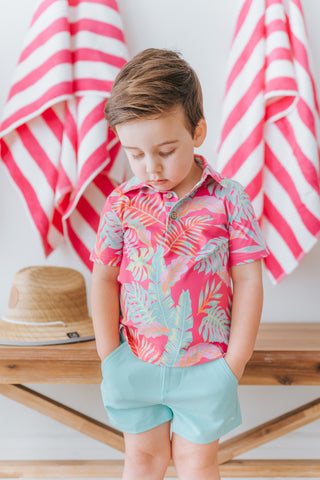 Child wearing a colorful tropical shirt and light blue shorts standing in front of pink and white striped towels.