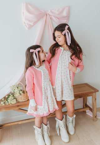 Two young girls in matching outfits with pink bows standing in a room with a large pink bow decoration.