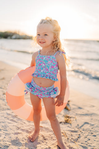 Young girl in a floral swimsuit standing on a beach with a pink inflatable ring.