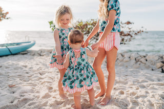 Three children in matching outfits standing on a beach with kayaks in the background.