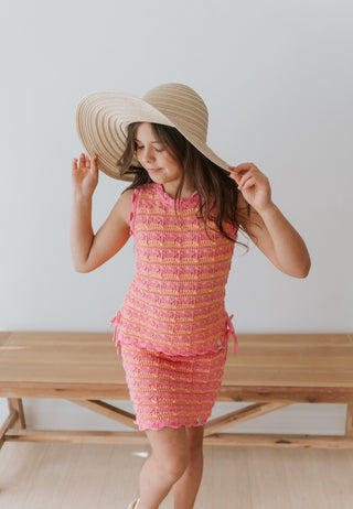 Young girl wearing a pink dress and large straw hat indoors.