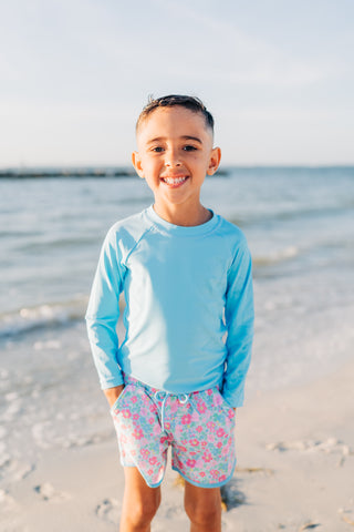 Child wearing a blue long-sleeve shirt and floral shorts standing on a beach.