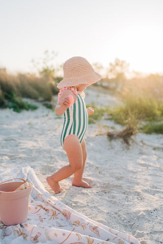 Child in a striped swimsuit and sun hat on a sandy beach with a pink bucket and towel.