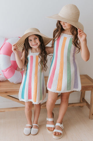 Two children wearing matching rainbow-striped dresses and wide-brimmed hats indoors.