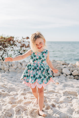 Child in a floral dress standing on a sandy beach with ocean and rocks in the background