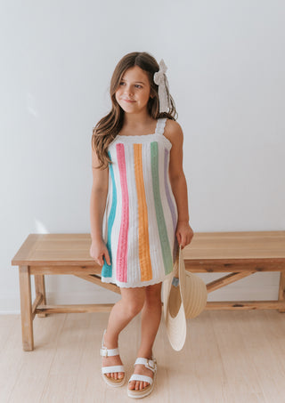 Young girl wearing a colorful striped dress standing in a room with a wooden bench.